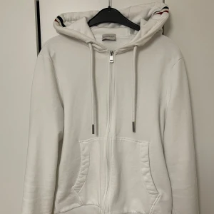 Vit hoodie från moncler - Säljer en stilren vit hoodie från Moncler med dragkedja och randiga detaljer i rött och blått på huvan och ärmarna. Perfekt för en avslappnad look. Den har en justerbar huva med snören och praktiska fickor framtill. Inte äkta. Den är i storlek M. Kvitto finns inte!