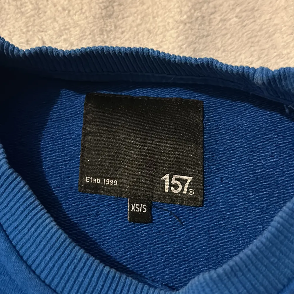 Säljer en blå tröja från 157 i storlek XS/S. Tröjan har långa ärmar och en rund halsringning med ribbade muddar vid ärmslut och nederkant. Perfekt för en avslappnad stil.. Neuletakit & Villapaidat.
