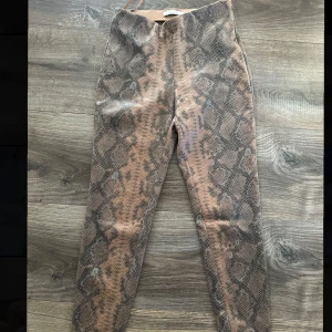 Ormmönstrade byxor från Vila - Säljer ett par coola ormmönstrade byxor från Vila. De är i ett snyggt brunt och svart mönster som verkligen sticker ut. Perfekta för en trendig look, oavsett om det är till vardags eller fest. Byxorna har en tight passform och är gjorda i ett bekvämt material.