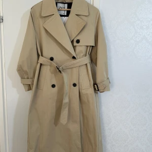 Beige trenchcoat i bomull från Zara - Säljer en snygg beige trenchcoat från Zara i nytt skick. Den har en klassisk design med dubbelknäppning och ett bälte i midjan. Perfekt för höst och vår! Långärmad med stiliga detaljer som axelklaffar och en slits baktill. Passar både till vardags och lite finare tillfällen.