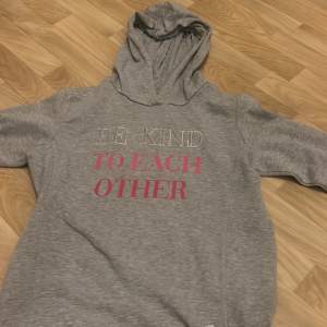 Mysig grå hoodie med texten 'BE KIND TO EACH OTHER' i vitt och rosa. Perfekt för en avslappnad stil. Den har långa ärmar och en bekväm huva. Passar bra till både jeans och mjukisbyxor. Perfekt för höst och vår!