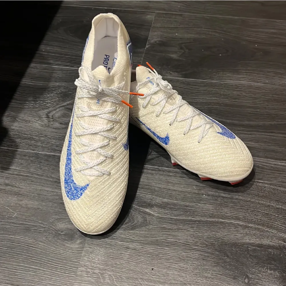 Snygga vita fotbollsskor från Nike med blå glittriga detaljer och orangea dobbar. Skorna har en modern design med snörning och är perfekta för fotbollsträning eller match. De har en tight passform för optimal kontroll på planen. . Skor.