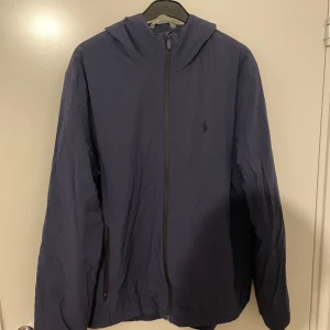Mörkblå vindjacka med huva - Säljer en mörkblå vindjacka i bra skick. Den har en praktisk huva och dragkedja framtill. Perfekt för höst- och vårväder. Jackan har en enkel och stilren design, vilket gör den lätt att matcha med olika outfits.