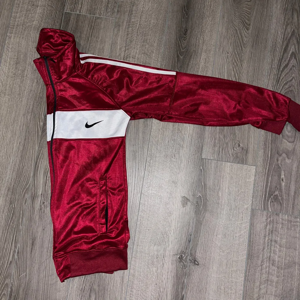 Säljer en röd träningsjacka från Nike med vita och svarta detaljer. Jackan har en dragkedja framtill och långa ärmar med vita ränder längs axlarna. Perfekt för träning eller en sportig look. Den är i bra skick och redo för nya äventyr! 🏃‍♂️. Jackor.