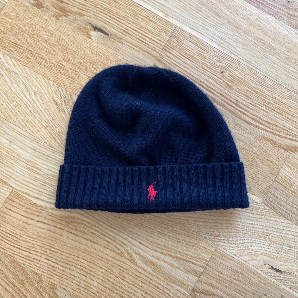 Snygg mörkblå mössa från Polo Ralph Lauren med röd broderad logga framtill. Den är stickad och har en ribbad kant som ger en skön passform. Perfekt för kyliga dagar och en stilren look. 🖤. Övrigt.