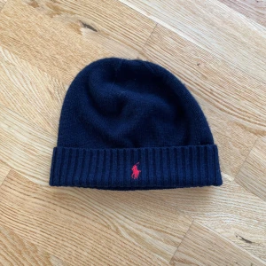 Mörkblå  mössa från Polo Ralph Lauren - Snygg mörkblå mössa från Polo Ralph Lauren med röd broderad logga framtill. Den är stickad och har en ribbad kant som ger en skön passform. Perfekt för kyliga dagar och en stilren look. 🖤