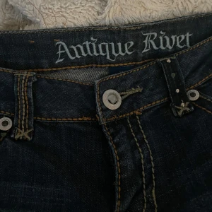 Mörkblå low rise jeans från Antique Rivet - Snygga mörkblå jeans från Antique Rivet med coola detaljer. De har dekorativa stenar på bakfickorna och en broderad design. Några stenar saknas på fickorna annars är dom i fint skick. 