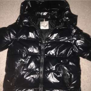 Svart pufferjacka från Moncler - Säljer en snygg svart pufferjacka från Moncler i mycket bra skick. Jackan har en glansig finish och dragkedja framtill. Perfekt för kalla dagar med sin varma vaddering. Den har också en cool detalj med en serieteckning på insidan. Passar perfekt för vintern!