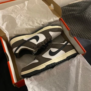Nike Dunk i brunt och vitt - Säljer ett par Nike Dunk i brunt och vitt. Skorna är i nytt skick och kommer i originalkartong. De har en snygg design med bruna detaljer och vit bas, samt klassisk Nike-logga på sidan. Perfekta för både vardag och fest!