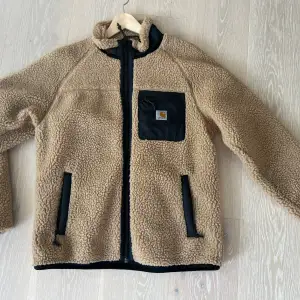 Säljer en supermysig beige teddyjacka från Carhartt i storlek M. Jackan har en svart dragkedja och en praktisk bröstficka med Carhartt-logga. Perfekt för höst och vinter, håller dig varm och stilren. Passar både till vardags och chilliga utflykter. 🧥✨