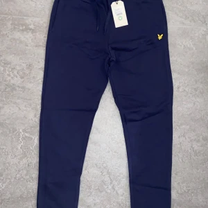 Mörkblå mjukisbyxor med gul logga - Säljer ett par mörkblå mjukisbyxor från Lyle & Scott i nytt skick. De har en gul broderad logga på ena benet och snörning i midjan för perfekt passform. Perfekta för en avslappnad dag hemma eller en snabb tur till affären. Superbekväma och stilrena! Och även inte använda i plast paket