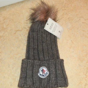 Brun stickad mössa från Moncler - Säljer en superfin brun stickad mössa från Moncler med en fluffig pälsboll på toppen. Den har en ribbad design och Monclers logga broderad framtill. Perfekt för att hålla sig varm och snygg under kalla dagar! 🧣✨