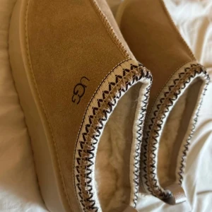 Beige mocka tofflor från UGG - Mysiga beige tofflor från UGG i mjuk mocka med fluffigt foder. Perfekta för kalla dagar hemma. De har en snygg stickad kantdetalj och en bekväm passform.  Storlek 37! Aldrig använt💕