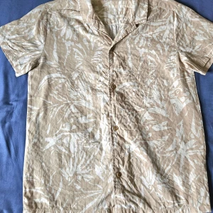 Beige kortärmad skjorta från Kappahl - Snygg beige skjorta från Kappahl med korta ärmar och ett diskret blommönster. Perfekt för varma dagar med sin lätta och luftiga design. Skjortan har knappar framtill och en klassisk krage. Använd 1 tillfälle.