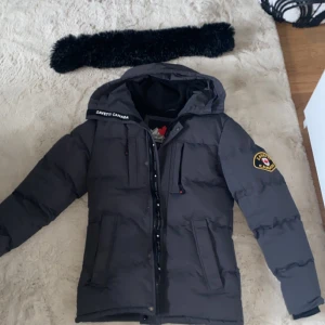 Grå Zavetti Canada jacka - Säljer en grå dunjacka från Zavetti Canada med avtagbar fuskpäls på huvan. Jackan har en dragkedja och knappar framtill samt flera praktiska fickor. Perfekt för kalla dagar med sin varma och vadderade design.