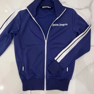 Palm angels tröja - Säljer en snygg blå track jacket från Palm Angels med vita ränder längs ärmarna och dragkedja framtill. Tröjan har en hög krage och två fickor med dragkedja. Tröjan är i ett mycket bra skick och säljer den för att jag inte längre använder den.