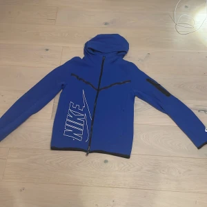 Blå hoodie från Nike - Säljer en blå hoodie från Nike med dragkedja och stor logga på sidan. Den har en huva och långa ärmar, perfekt för en sportig look. Passar bra för träning eller som en casual jacka.