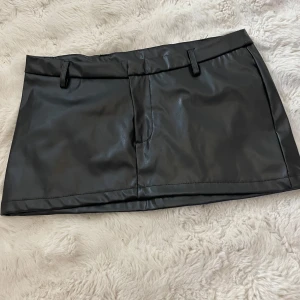 Svart lågmidjad mini kjol i skinn  - Snygg svart kortkjol i skinnimitation med bälteshällor och fickdetaljer baktill. 🤍