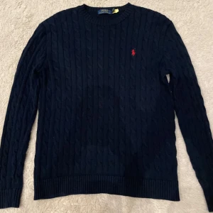 Mörkblå stickad tröja från Polo Ralph Lauren - Tröjan passar även xs. Säljer en snygg mörkblå stickad tröja från Polo Ralph Lauren med det klassiska röda logotypbroderiet på bröstet. Tröjan har ett kabelstickat mönster och långa ärmar, perfekt för kyligare dagar. Den är i mycket bra skick.