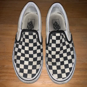 Svartvita slip-on sneakers från Vans - Klassiska slip-on sneakers från Vans med ett ikoniskt svartvitt rutigt mönster. Skorna har en vit sula och är perfekta för en avslappnad stil. 