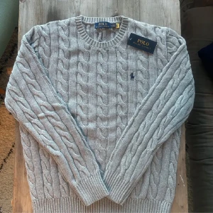 Grå kabelstickad tröja från Polo Ralph Lauren - Säljer en stilren grå kabelstickad tröja från Polo Ralph Lauren. Tröjan har långa ärmar och en klassisk rund halsringning. Perfekt för kyligare dagar och ger en tidlös look.
