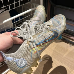 Silvriga fotbollsskor med färgglada detaljer - Snygga silvriga fotbollsskor med färgglada detaljer i blått, gult och rött. Skorna har snörning och en modern design som passar perfekt för fotbollsplanen.