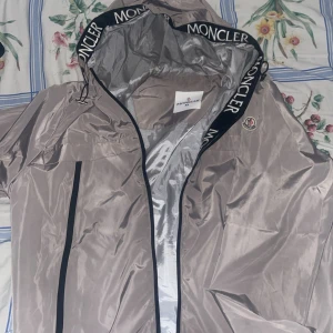 Beige vindjacka från Moncler - Snygg beige vindjacka från Moncler med svarta dragkedjor och logotypdetaljer på huvan. Storleken är XL men jag använder L och den sitter bra. Jackan har en stilren design med en lätt glansig yta och är perfekt för blåsiga dagar. Den har en praktisk dragkedja framtill och en lång fick. Den är också gjord för regn.