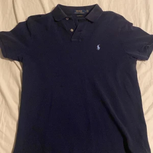 Mörkblå pikétröja från Ralph Lauren - Säljer en stilren mörkblå pikétröja från Ralph Lauren i custom slim fit. Tröjan har en klassisk krage med knappar och det ikoniska broderade logotypen på bröstet. Perfekt för en avslappnad men ändå elegant look.