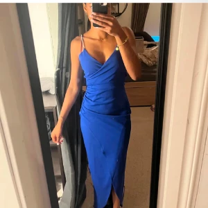 Blå klänning från Quiz - Snygg blå klänning från Quiz med smala axelband och omlott-design. Klänningen har en elegant slits framtill och en figurnära passform som framhäver silhuetten. Perfekt för speciella tillfällen!