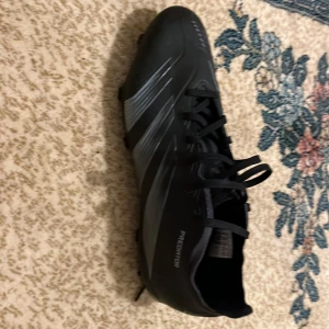 Svarta fotbollsskor Adidas Predator - Säljer ett par svarta Adidas Predator fotbollsskor med snörning. Skorna har en stilren design med diskreta ränder och är perfekta för fotbollsplanen. Tillverkade i syntetmaterial för hållbarhet och prestanda. Utan boxen!