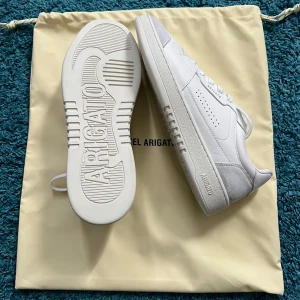 Vita sneakers från Axel Arigato - Snygga vita sneakers från Axel Arigato med stilren design. Skorna har en vit sula och detaljer i grå mocka. Perfekta för en minimalistisk look.