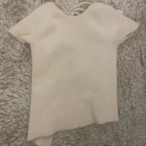 Beige stickad topp med snörning - Säljer en beige stickad topp från Zara med korta ärmar och snygg snörning i ryggen. Perfekt för en stilren look. Toppen har en asymmetrisk nederkant som ger en unik touch.