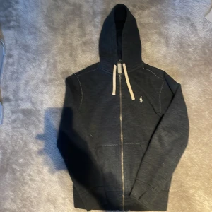 Mörkblå hoodie från Polo Ralph Lauren - Säljer en snygg mörkblå hoodie från Polo Ralph Lauren med dragkedja och huva. Tröjan har vita snören och en liten broderad logga på bröstet. Perfekt för en avslappnad stil. Pris går diskutera 