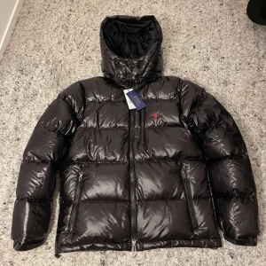 Svart pufferjacka från Polo Ralph Lauren - Säljer en svart pufferjacka från Polo Ralph Lauren med avtagbar huva och dragkedja framtill. Jackan har en glansig yta och två fickor med dragkedja. Perfekt för kyliga dagar!