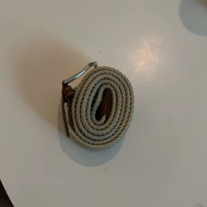Beige flätat bälte med silverfärgat spänne - Säljer ett snyggt beige flätat bälte med ett stilrent silverfärgat spänne. Perfekt för att ge en extra touch till din outfit. Bältet har bruna detaljer vid spännet som ger en fin kontrast.100cm långt bälte