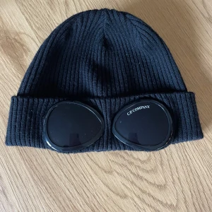 Svart ribbad mössa från C.P. Company - Snygg svart ribbad mössa från C.P. Company med inbyggda glasögon på framsidan. Perfekt för att ge en unik touch till din outfit. Mössan är tillverkad i ett mjukt och bekvämt material.