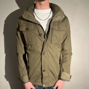 Olivgrön Field jacka - Snygg olivgrön field jacka med knappar och dragkedja från j.linberg. Jackan har långa ärmar och flera praktiska fickor framtill. Perfekt för en stilren look. Skriv vid intresse 🤩