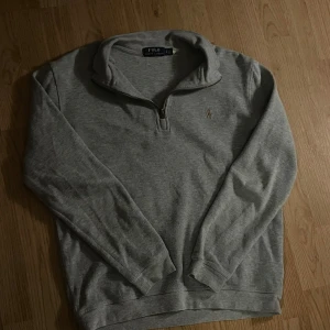 Grå tröja med dragkedja från Ralph Lauren st S - Säljer en stilren grå tröja från Ralph Lauren med halv dragkedja och broderad logga på bröstet. Perfekt för en avslappnad och snygg look. Tröjan har långa ärmar och är tillverkad i ett mjukt material. St S