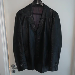 Skinnjacka från Monza - Snygg svart skinnjacka från Monza med klassisk design. Jackan har en stilren krage och knappar framtill. Insidan är fodrad med ett mönstrat tyg i röd och blå nyans. Perfekt för en cool och tidlös look.