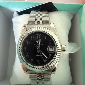 Silverfärgad klocka för kvinnor  - Elegant silverfärgad klocka med svart urtavla och datumvisning. Klockan har en klassisk länkarmband och arabiska siffror på urtavlan. Perfekt för den som söker en stilren och tidlös accessoar. Klockan är inte tjock, och det är för stt den är för kvinnor. 