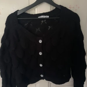 Svart stickad kofta från Nakd - Säljer en svart stickad kofta från Nakd med dekorativa knappar framtill. Koftan har ett fint mönster och långa ärmar, perfekt för kyligare dagar. Den är både stilren och bekväm att bära.