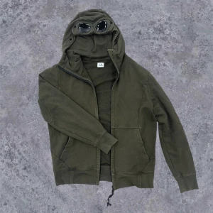 C.P. Company goggle zip-up - Säljer en grön zip-up från C.P. Company med inbyggda goggles i huvan 🥽Använt en del, finns två små färgfläckar som knappt syns men de borde gå att ta bort 😶‍🌫️Sitter lite baggy på mig som brukar ha storlek small, så funkar för S-M 🤙Pris kan diskuteras‼️