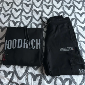Svart hoodie och byxor Hoodrich - Säljer en svart hoodie och matchande byxor från Hoodrich. Hoodien har en stor logga på bröstet och ryggen, samt en känguruficka framtill. Byxorna har en logga på benet och en praktisk ficka. Perfekt för en avslappnad stil.