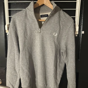 Grå tröja med dragkedja från Fred Perry - Säljer en stilren grå tröja från Fred Perry med halv dragkedja och broderad logga på bröstet. Perfekt för en avslappnad look. Tröjan har långa ärmar och ribbade muddar vid ärmslut och nederkant.