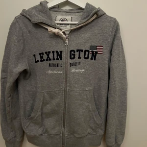 Grå hoodie från Lexington - Säljer en grå hoodie från Lexington med dragkedja och tryck framtill. Tröjan har en stor luva och praktiska fickor. 