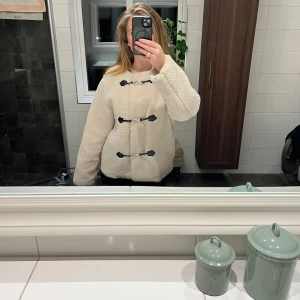 Beige teddyjacka från Gina Tricot - Mysig beige teddyjacka från Gina Tricot med svarta knäppdetaljer. Perfekt för kyliga dagar med sin fluffiga och varma design. Jackan har långa ärmar och en stilren look som passar till många olika outfits.