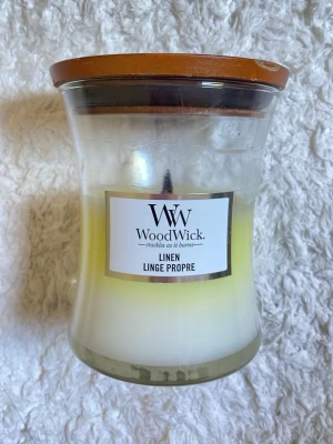 WoodWick doftljus Linen med trälock - Säljer ett WoodWick doftljus i doften Linen. Ljusen har en snygg glasburk med trälock och träveke som sprakar när det brinner. Färgen är vit med en lätt gul ton och etiketten är stilren. Perfekt för att skapa mysig stämning hemma.
