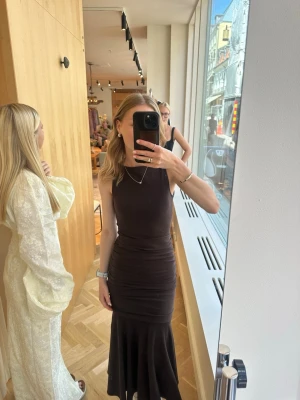 Mörkbrun långklänning med drapering - Snygg mörkbrun långklänning med breda axelband och draperad midja som ger en cool siluett. Klänningen är ärmlös och har en figurnära passform upptill med ett längre, flödande fall nedtill. Perfekt för dig som vill sticka ut. Slutsåld på Adoore 