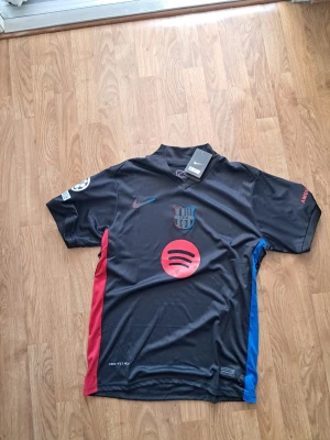Svart FC Barcelona matchtröja Nike - Säljer en svart FC Barcelona matchtröja från Nike med klubbmärke, Spotify-logga och färgdetaljer i rött och blått på sidorna. Tröjan har korta ärmar, tryck på både fram- och baksida samt officiella märken på ärmen. Perfekt för dig som älskar fotboll och Barca.