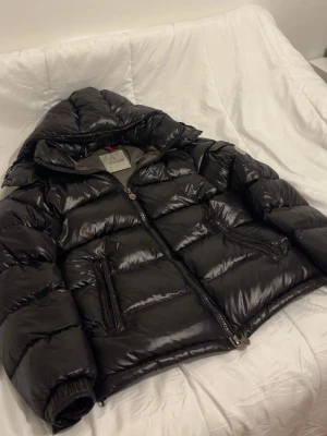 Svart pufferjacka från Moncler - Snygg svart pufferjacka från Moncler med glansig finish och huva. Jackan har två stora framfickor med dragkedja och klassisk Moncler-detalj på insidan. Perfekt för kalla vinterdagar och riktigt varm tack vare dunfyllning.